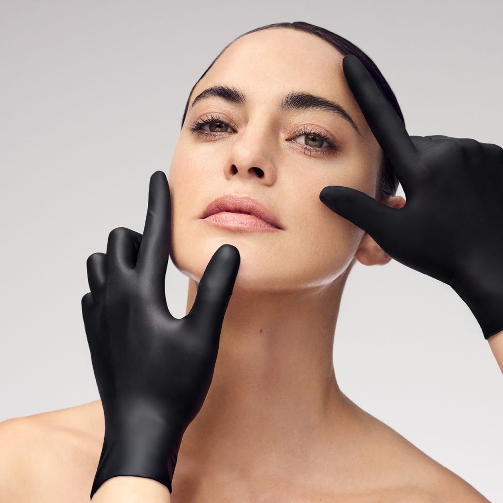 Gesicht einer Frau mit ebenmäßiger Haut, sanft umrahmt von zwei schwarz behandschuhten Händen – professionelle Kosmetik.